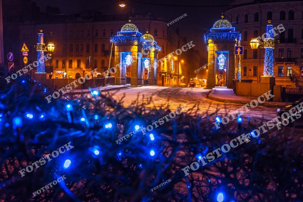 На фотографии изображен мост Ломоносова в Санкт-Петербурге, украшенный новогодними гирляндами и иллюминацией. Вечерняя атмосфера оживляется сиянием синего и золотистого света, подчеркивающего архитектурную красоту моста. Снежное покрытие улиц и яркие огни создают ощущение уюта и праздника. Это место особенно привлекательно зимой, когда Петербург преображается в сказочный город. Ломоносовский мост является одним из символов исторического центра города.